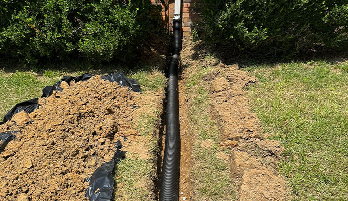 Exterior Drainage System Installation A trench in the yard with a black drainage pipe running through it